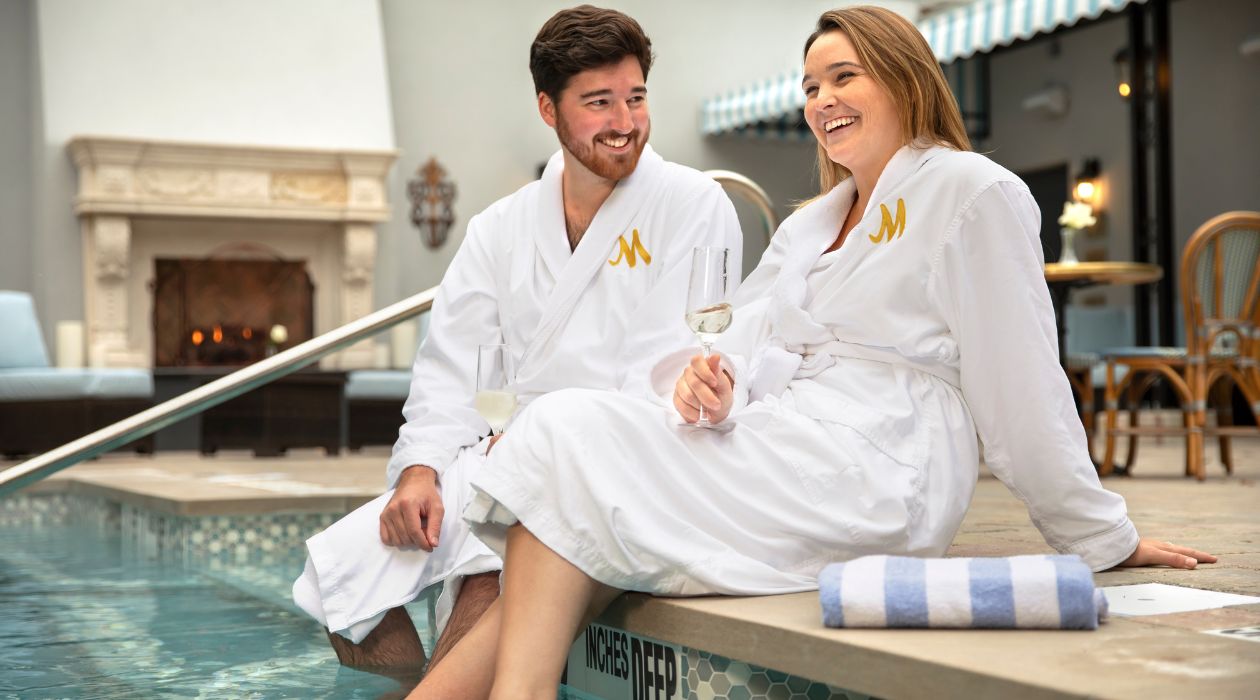 A man and woman relax by the pool wearing matching white robes and drinking glasses of wine at Mirbeau Inn and Spa in Rhinebeck.